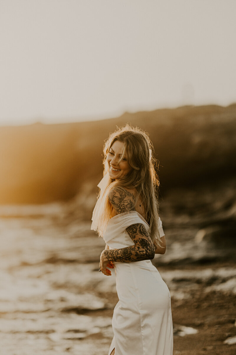 Femme en robe blanche souriante baignée par la lumière d'un coucher de soleil à la plage pendant une séance photo couple.