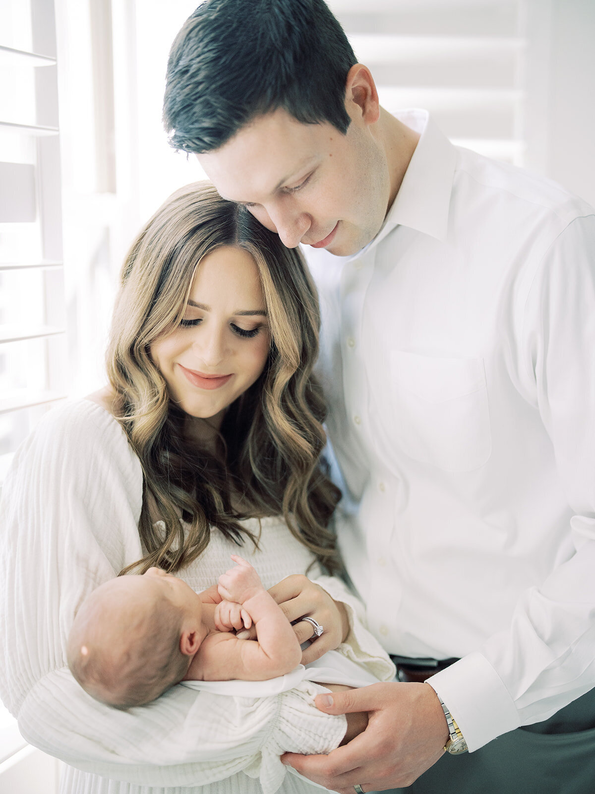 Brown-haired mother and father dressed in white stand huddled together holding their newborn baby girl during their Bethesda newborn session.