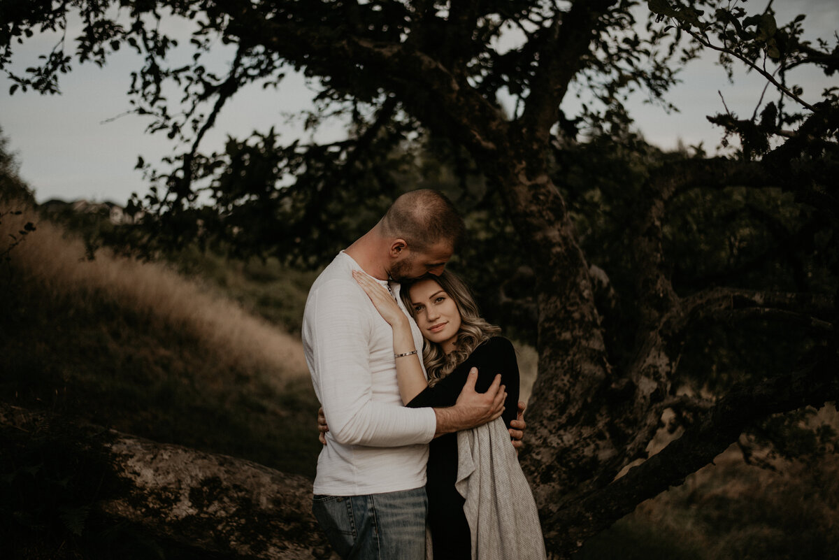 couple standing in front of a curved trunk tree. The guy is facing the girl and kissing her head, while she rests her head and hand against his chest. She is hugging him slightly sideways and looking right at the famera.
