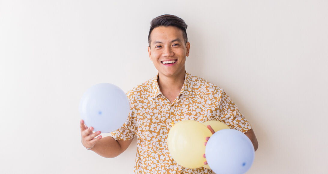 Sho Dewan, young Asian man, career coach and founder of Workhap is standing against a white wall, holding air balloons in his hands and laughing, looking straight at the camera