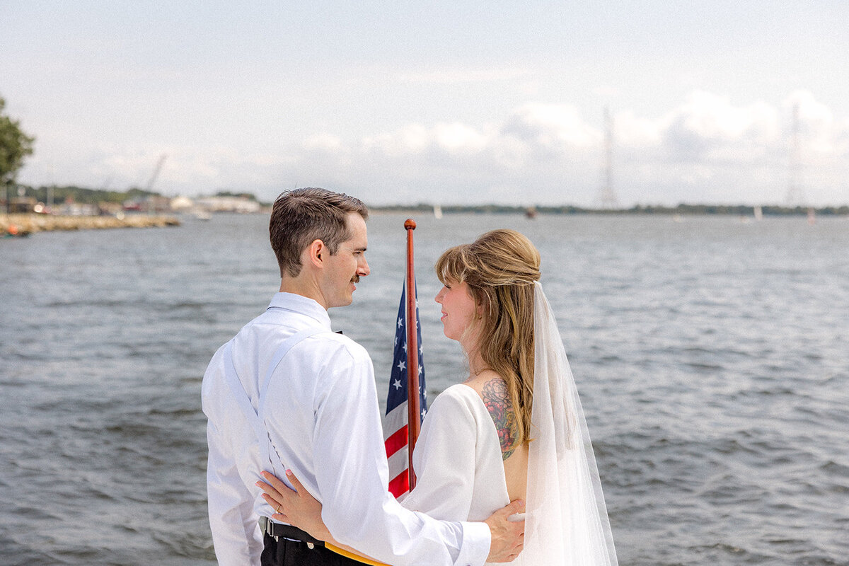 Bride and groom looking at each other with an american flag in between them and water in back