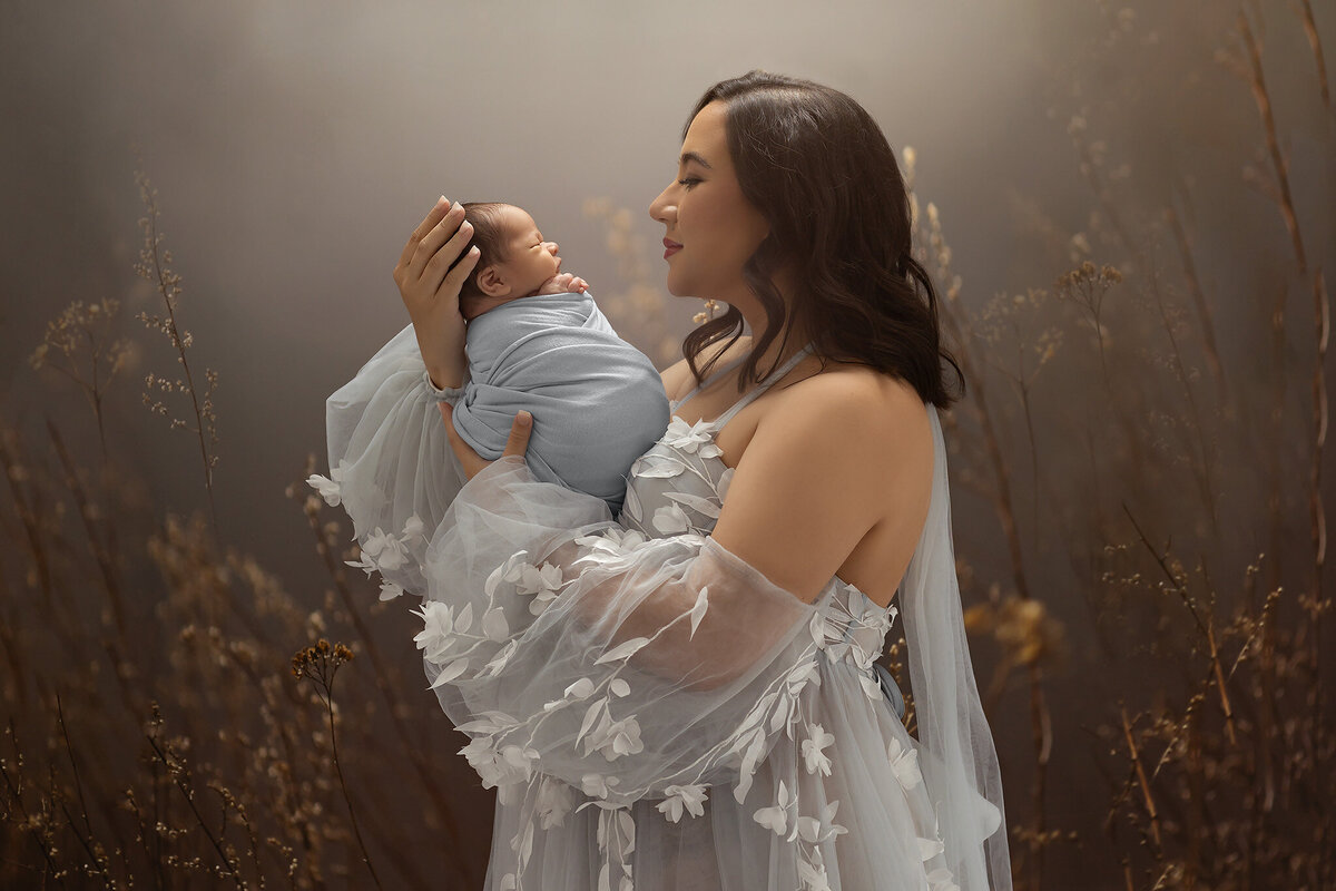Mother in a soft blue floral gown holding her swaddled newborn in a dreamy, nature-inspired studio setting with dried florals and gentle light, captured by Dallas newborn photographer Lindsay Walden
