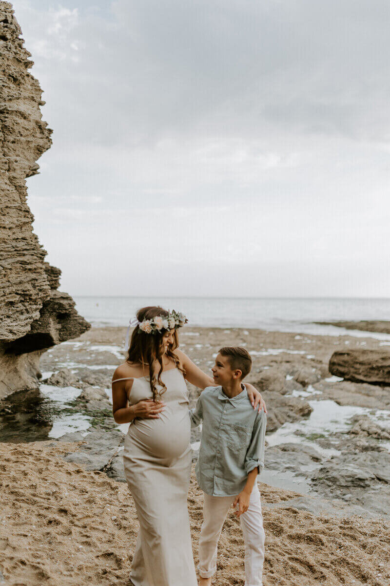 Maman enceinte dans une robe blanche, bras sur l'épaule de son grand garçon qui se tient à côté. Photo prise à la plage en vendée par Laura Termeau photographie.