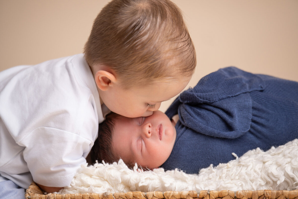 Young brother leans over to kiss his newborn brother for a family portrait studio