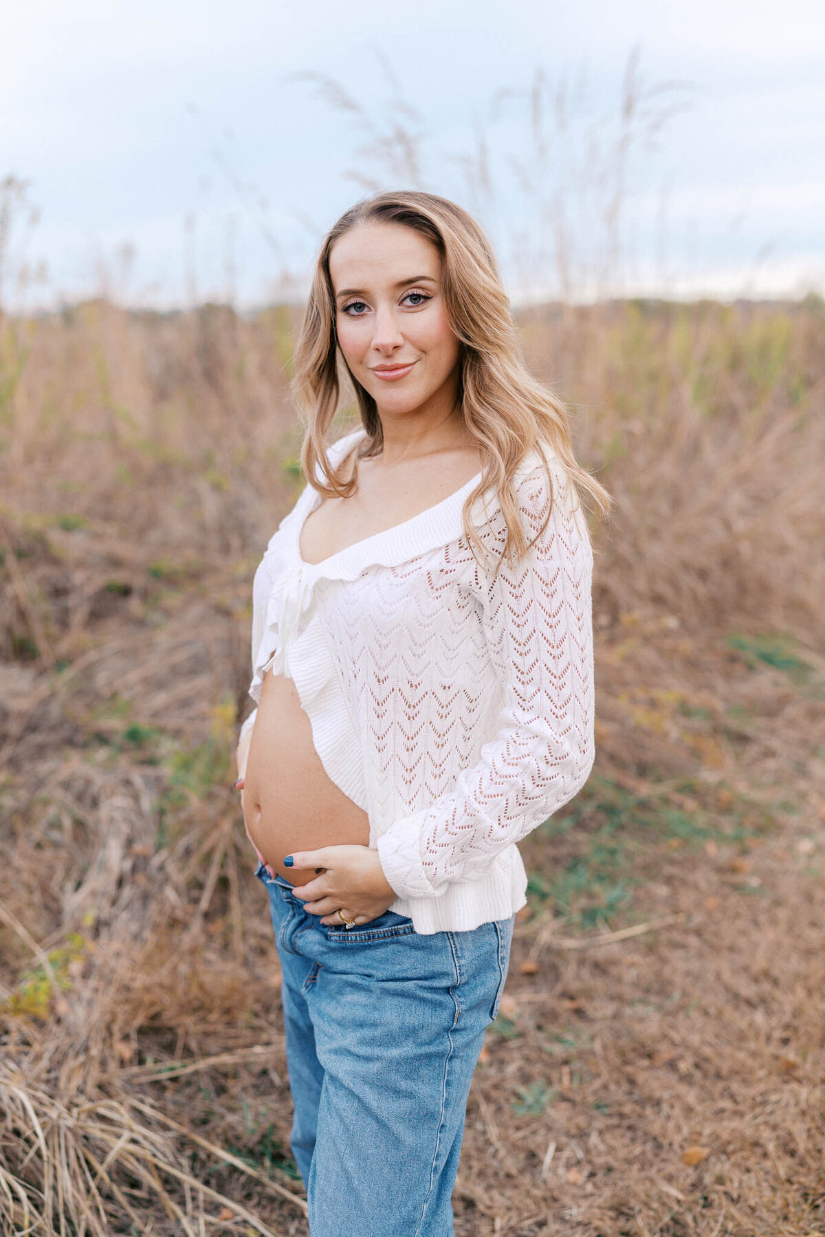 pregnant woman in white lace shirt holds belly in field during golden hour in knoxville tennessee
