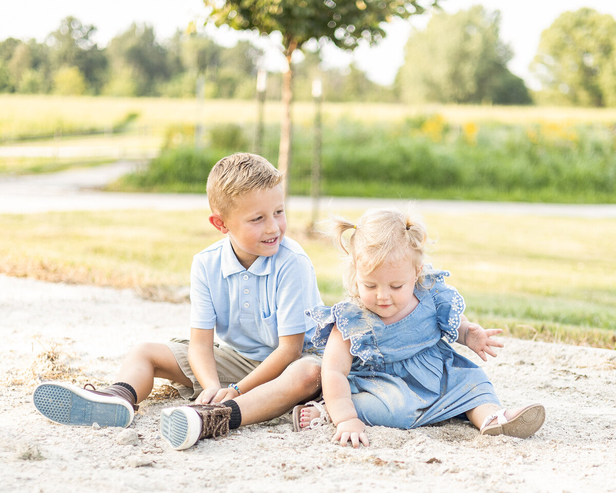 Young brother and sister sitting on the ground playing.  brother has short brown hair, wearing a partial button up short sleeve shirt, khaki shorts, brown shoes, smiling at his sister.  Sister has blonde hair in pigtails, wearing a blue dress, white sandals, and looking down at the ground