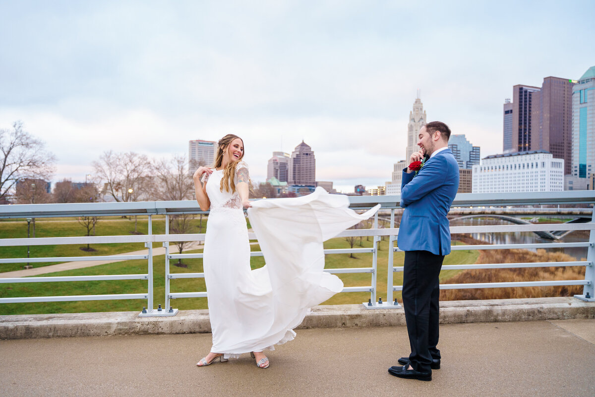 Bride swings her dress train as her groom checks her out in front of the Columbus skyline.