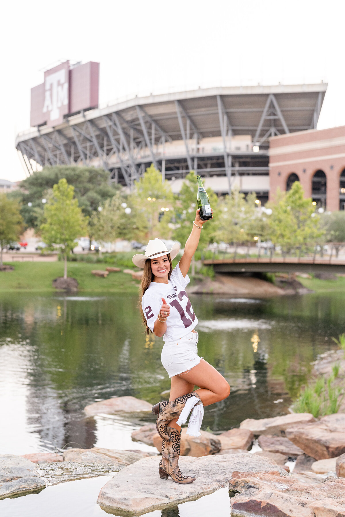 Texas A&M senior girl wearing white jersey and cowboy hat while smiling and holding up champagne bottle while on the rocks of Aggie Park in front of Kyle Field