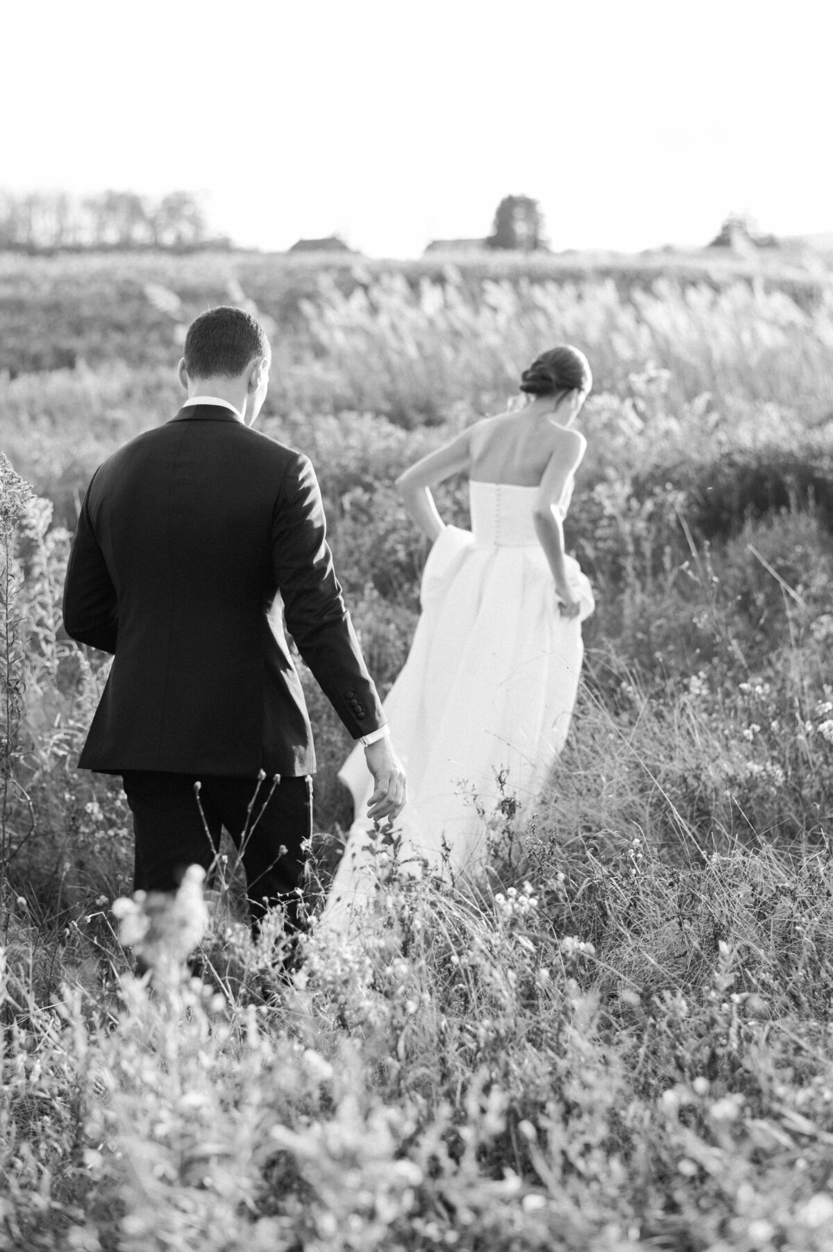 The groom follows the bride as she walks through the tall grass, her dress trailing behind her