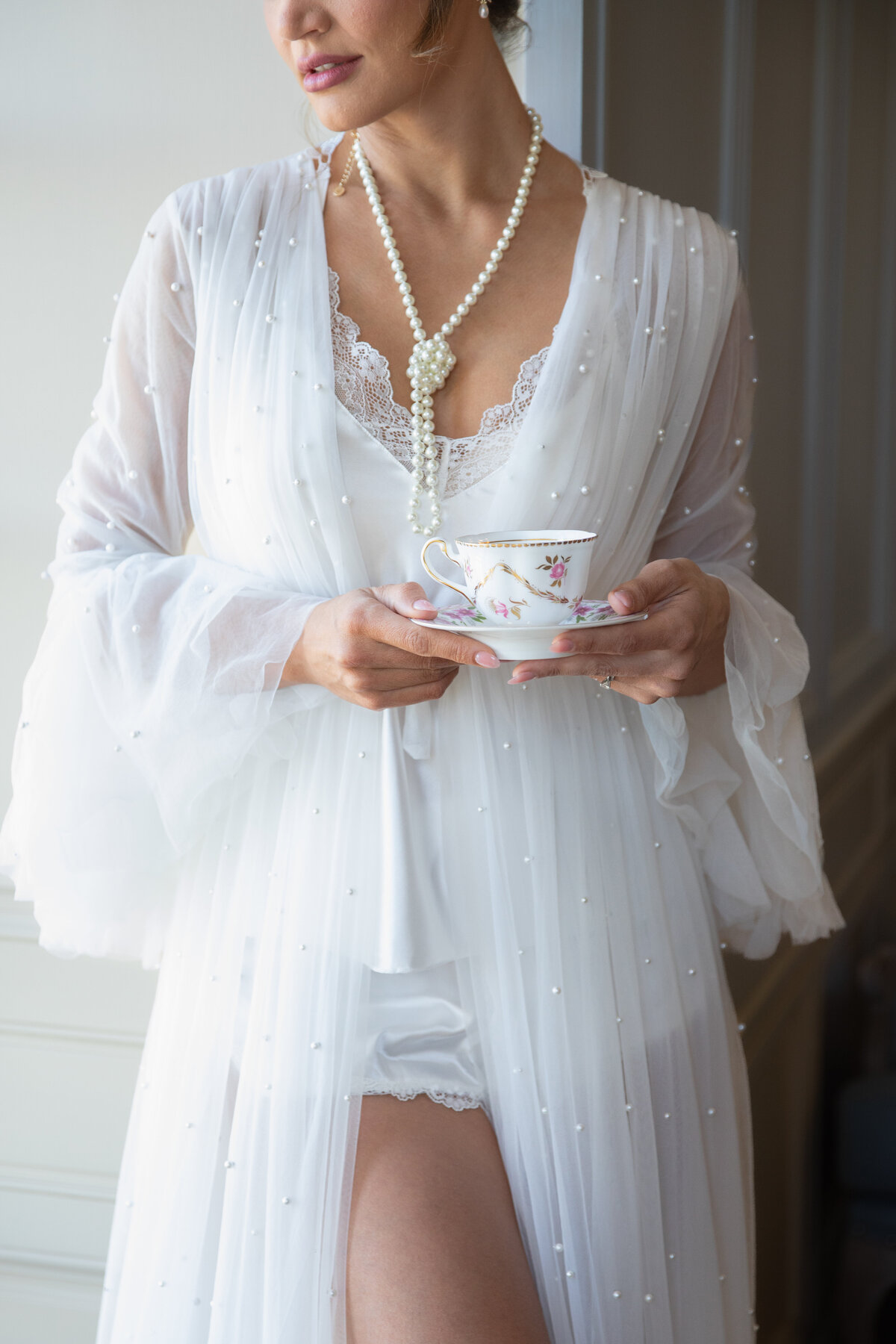 Wedding Photographers in RI | Kelsey Sheehan Photography Timeless Rhode Island Weddings | A person in a sheer, pearl-embellished white robe holds a floral teacup. They wear lace lingerie and a pearl necklace, exuding elegance and tranquility.