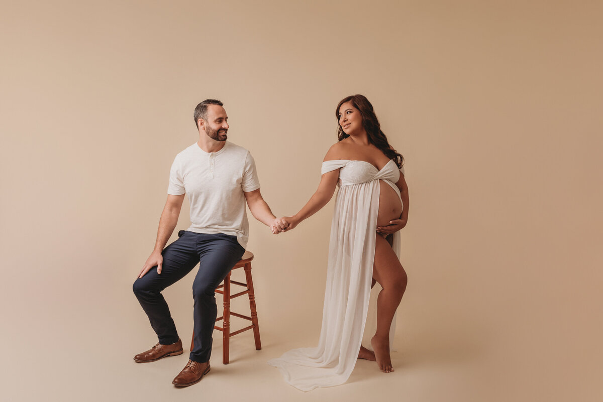 Pregnant woman at maternity and newborn studio of Atlanta posing for maternity portrait wearing white sheer maternity gown on tan backdrop holding hands with husband looking at eachother