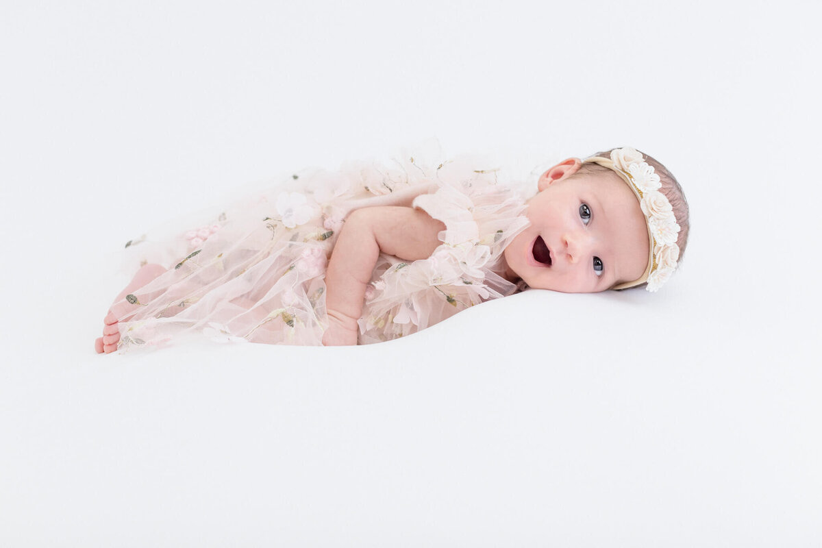 A baby wearing a floral headband and a frilly, lace dress lies on a white background. The baby is looking at the camera with a slight smile, wrapped in a soft, semi-transparent fabric.