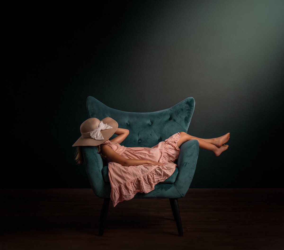 A little girl in an orange dress napping in a green chair with a hat over her eyes and light pouring in above her
