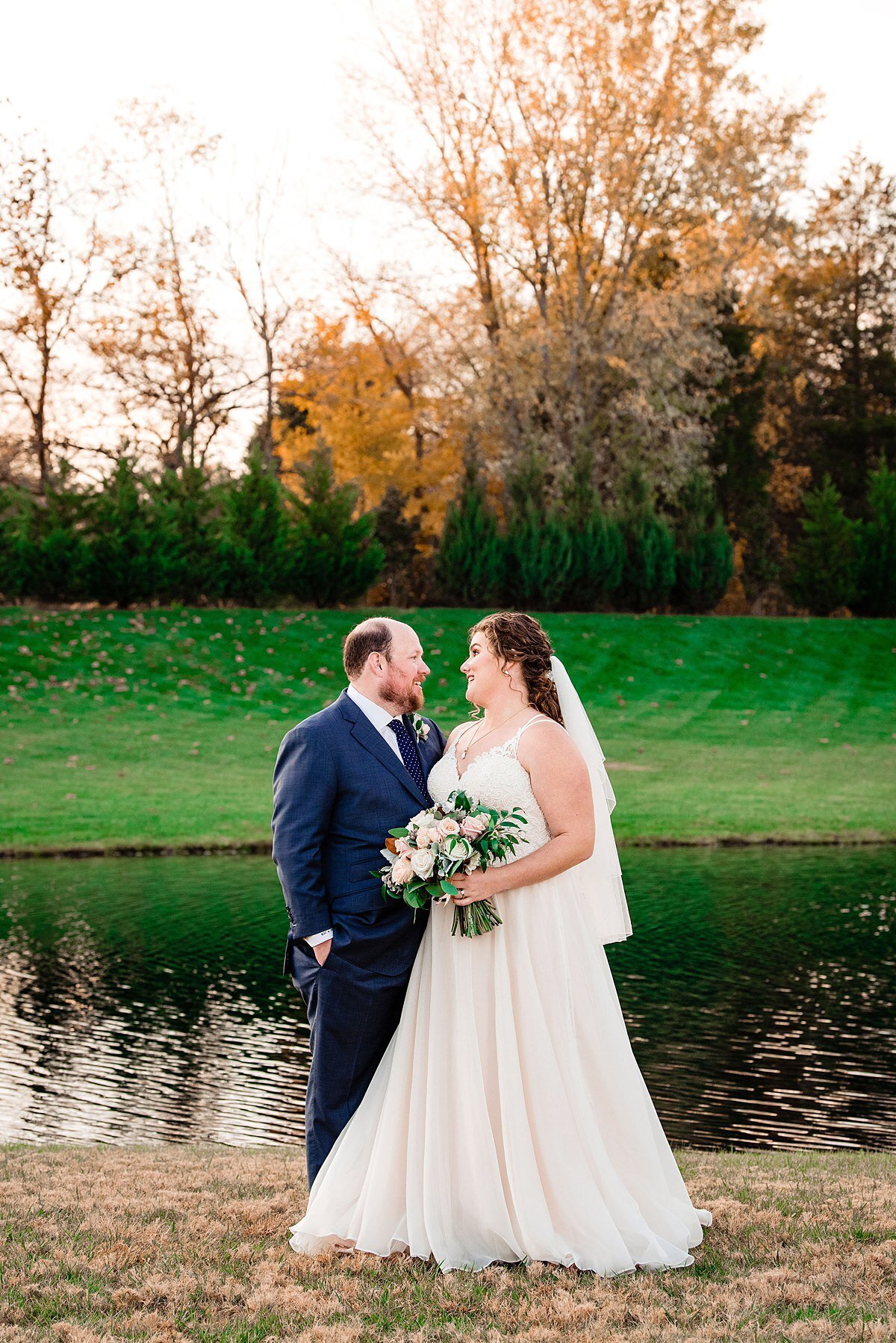 The bride wearing a lace v-neck a-line dress and long veil smiling back at her groom dressed in a navy blue suit and tie while holding a large round bouquet of ivory, white, blush, burgundy and soft gray flowers and greenery as they stand beside a lake at Sycamore Farms