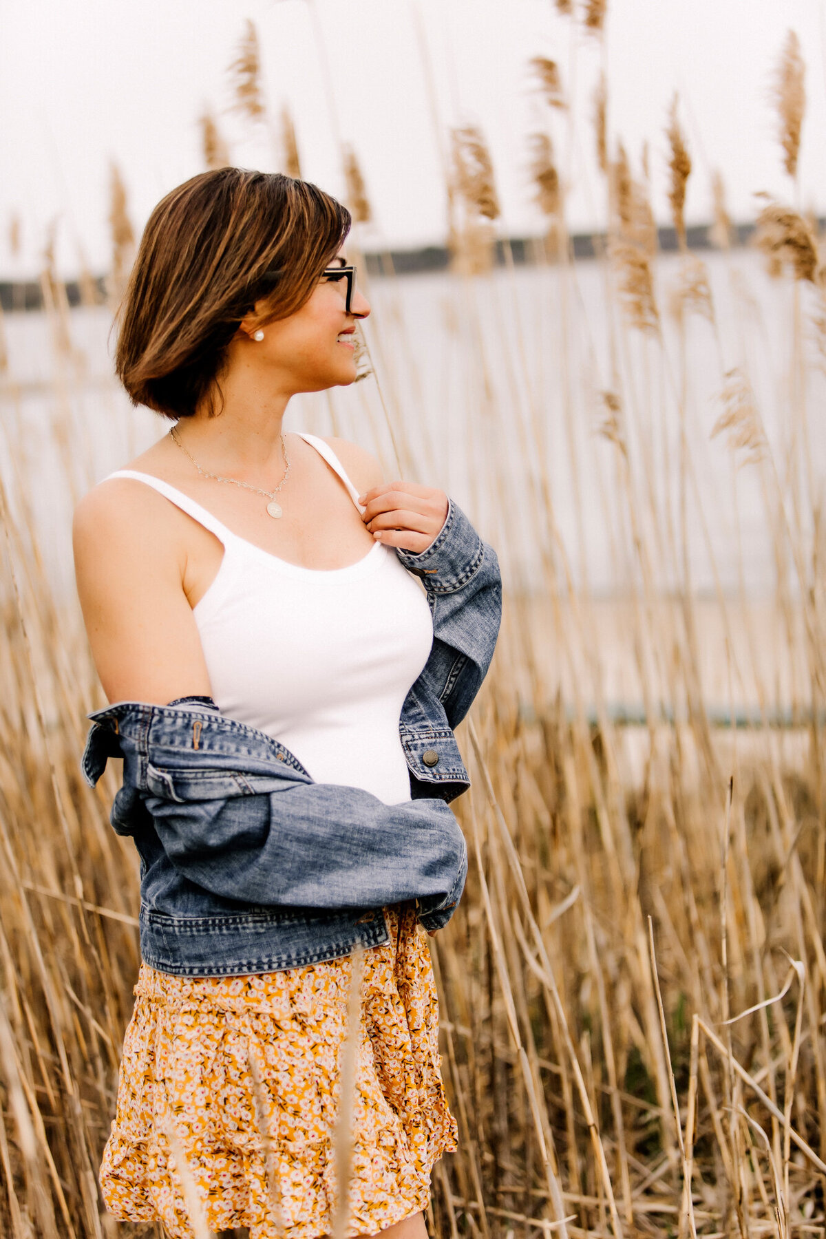 A female dating portrait client wearing a white tank top with a jean jacket off her shoulders, standing in front of tall grasses at the beach