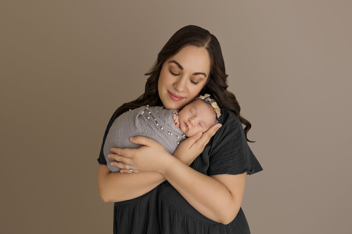 Mother lovingly holding her sleeping newborn baby wrapped in a grey blanket, standing against a neutral beige background.