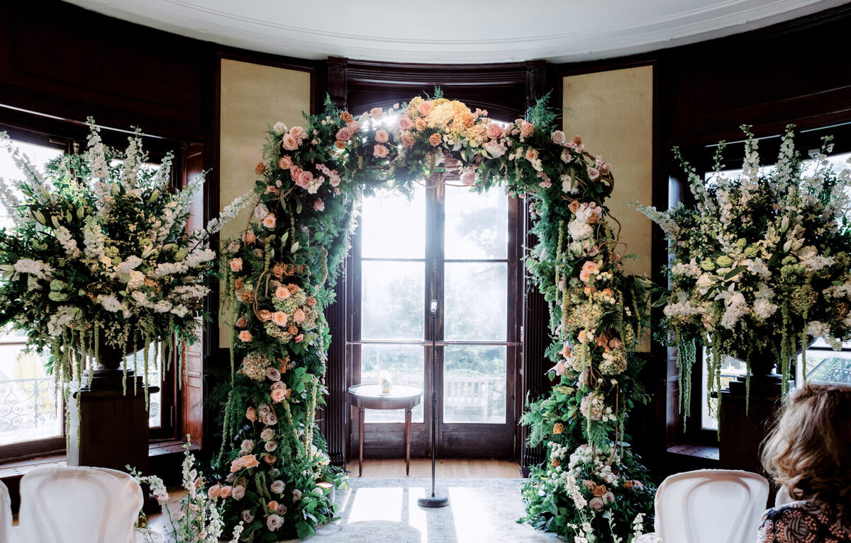 An arch with many flowers, inside a room, with large flower arrangements on both sides.