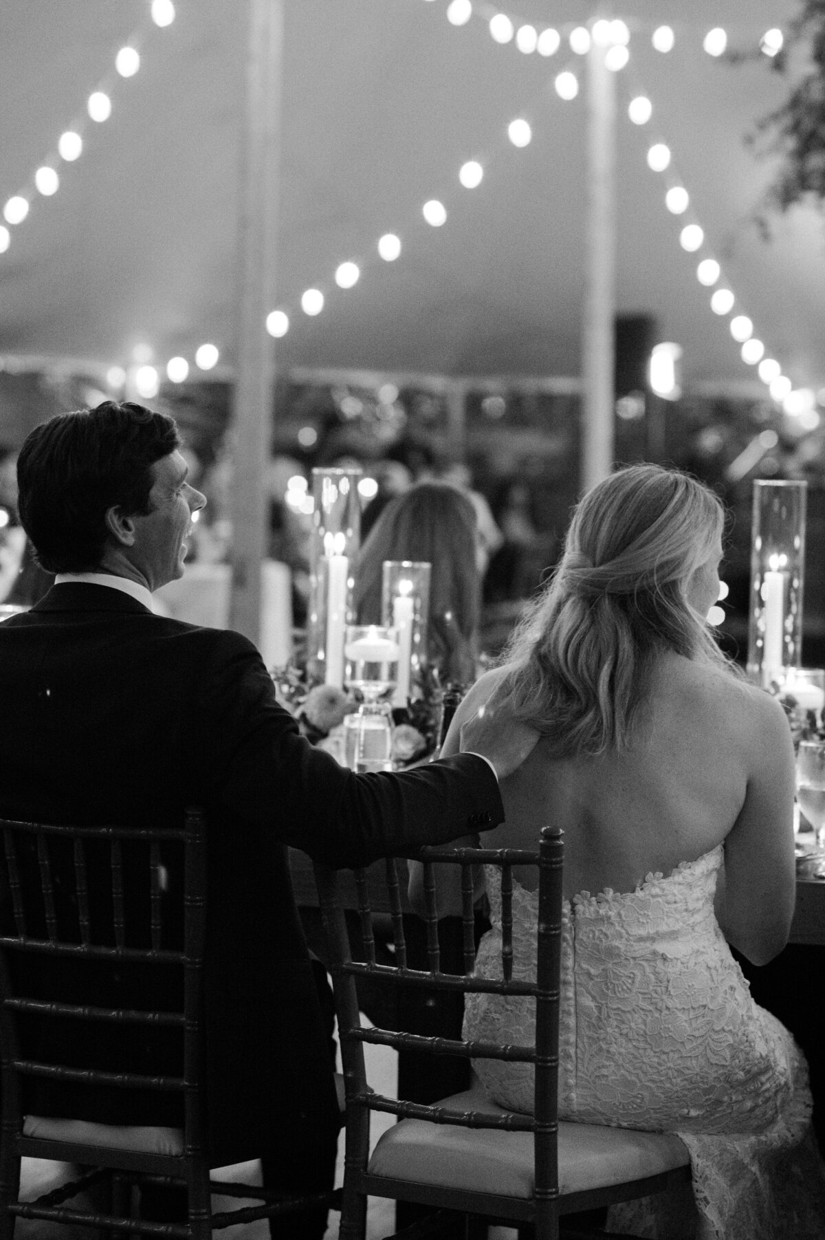 The groom gently places his hand on the bride's back as they sit at their reception under a tent filled with twinkling string lights