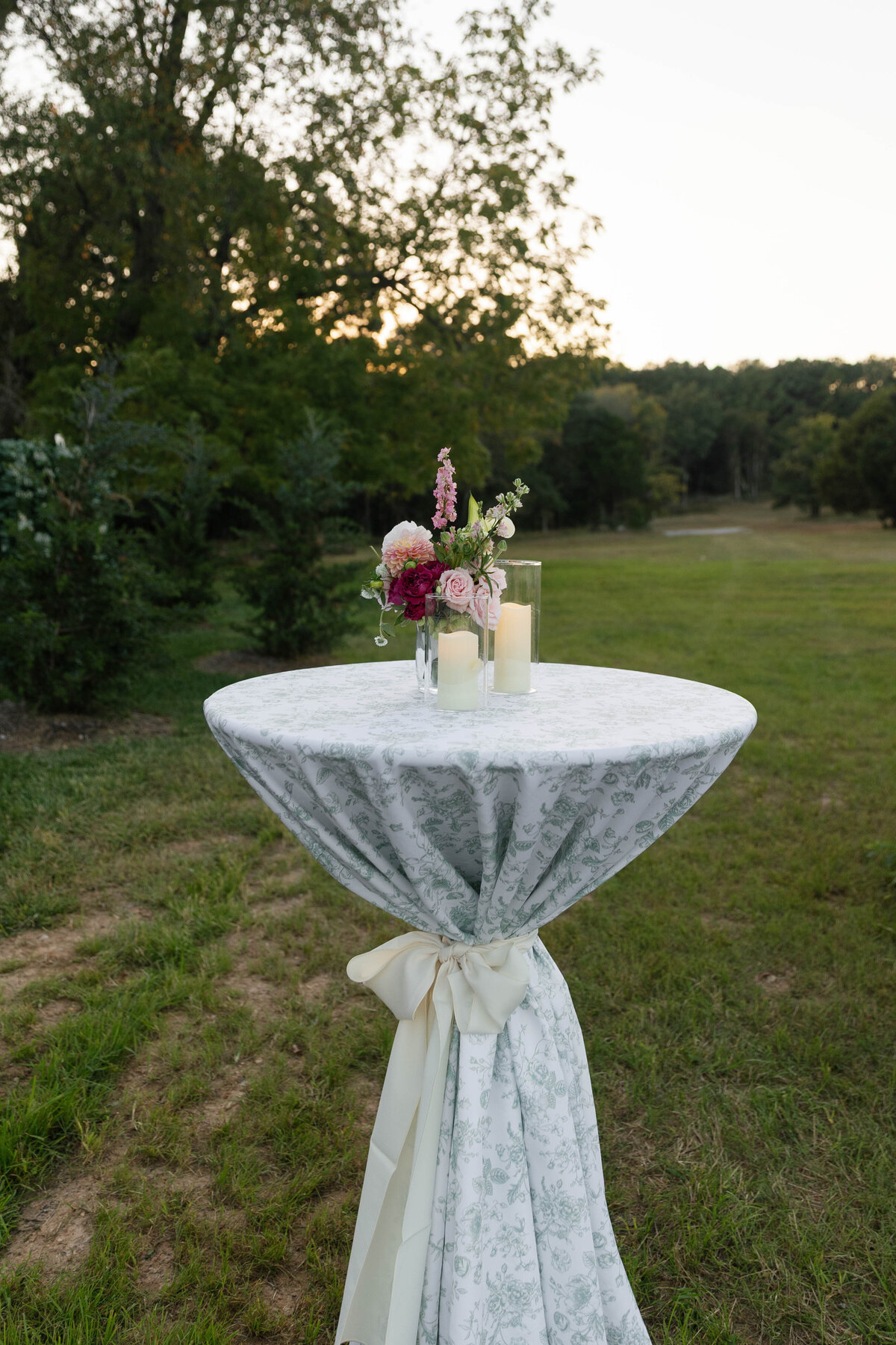 Tall cocktail table covered in a green toile linen and tied with an ivory sash at an outdoor Arkansas wedding reception. The table is decorated with a small arrangement of pink and burgundy flowers and glass-encased pillar candles, set against an open field and trees at sunset.