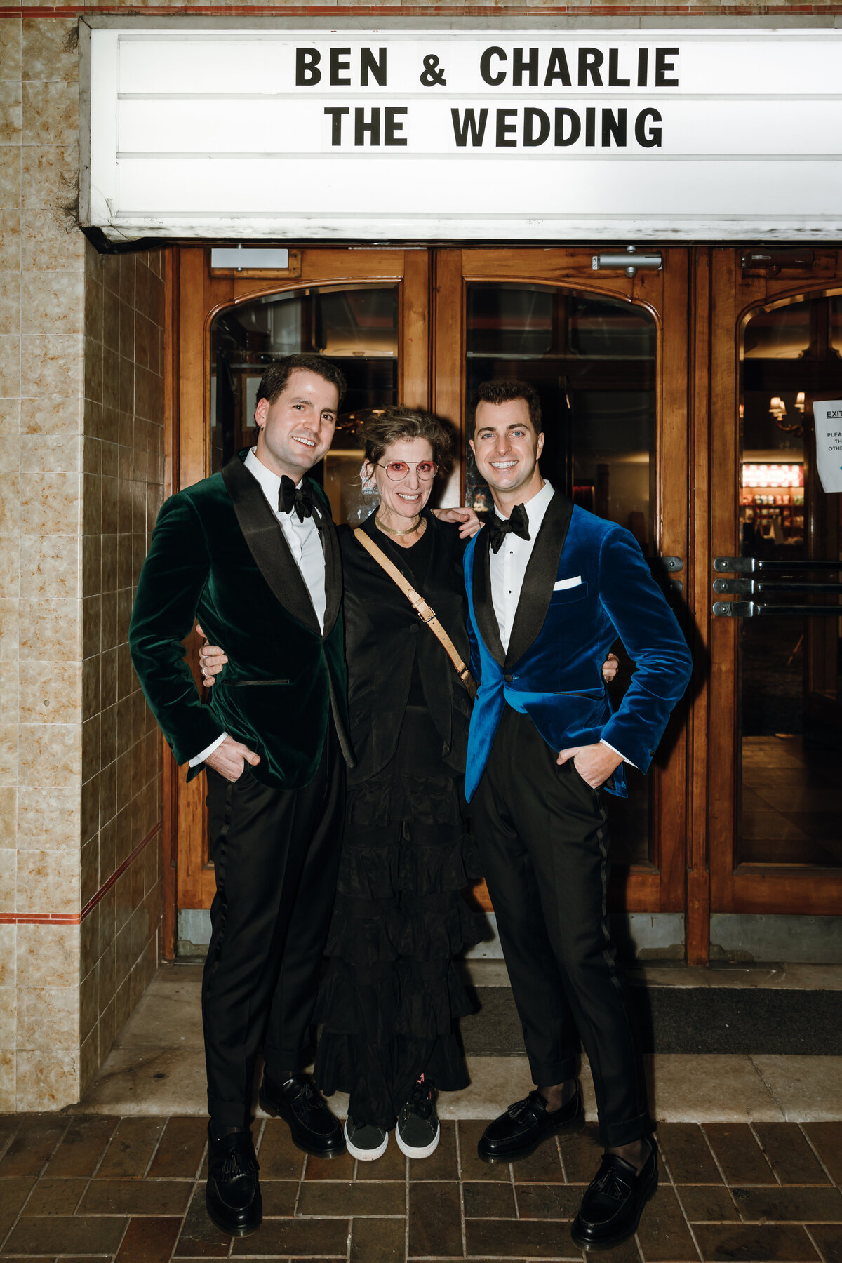 The grooms in green and blue velvet suits outside the entrance of regal cinema with a guest in between them.
