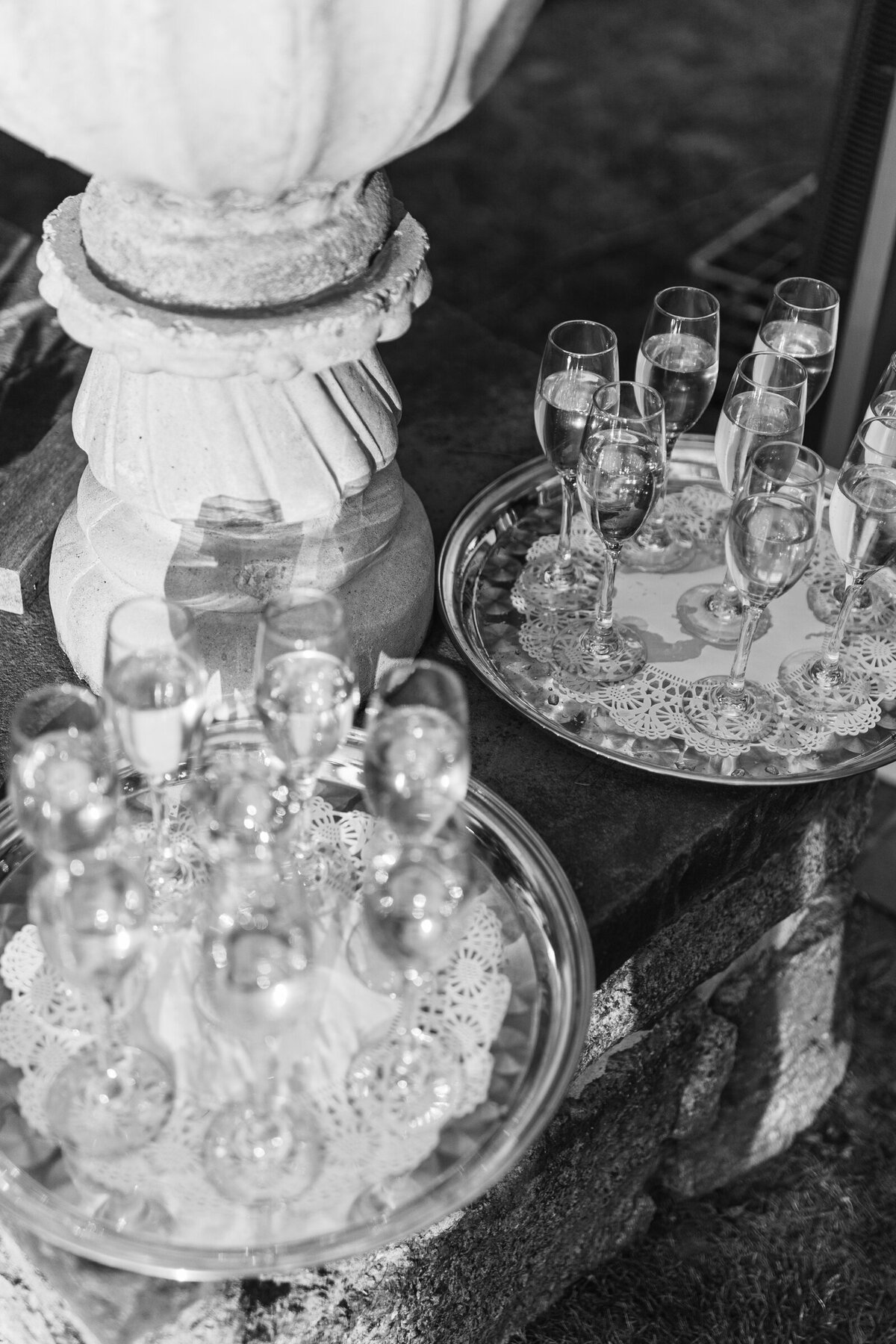 A black and white picture of champagne glasses on a silver tray, with harsh sunlight and heavy shadows. The image is editorial and moody.