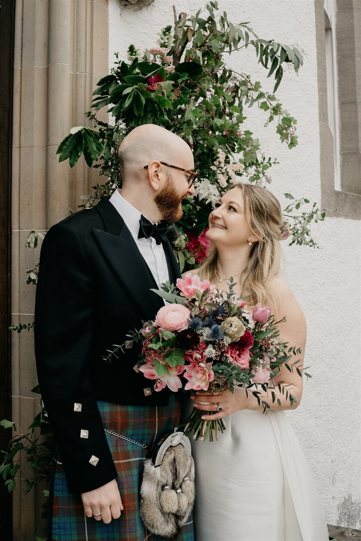 A bride and groom smile at each other as the groom wraps his arms around her.