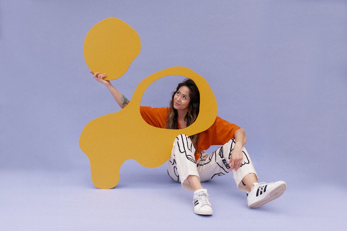 Woman sits on floor wearing an orange long sleeve and black and white abstract face printed pants and adidas in front of a purple backdrop while holding a wooden, mustard-colored abstract shape.