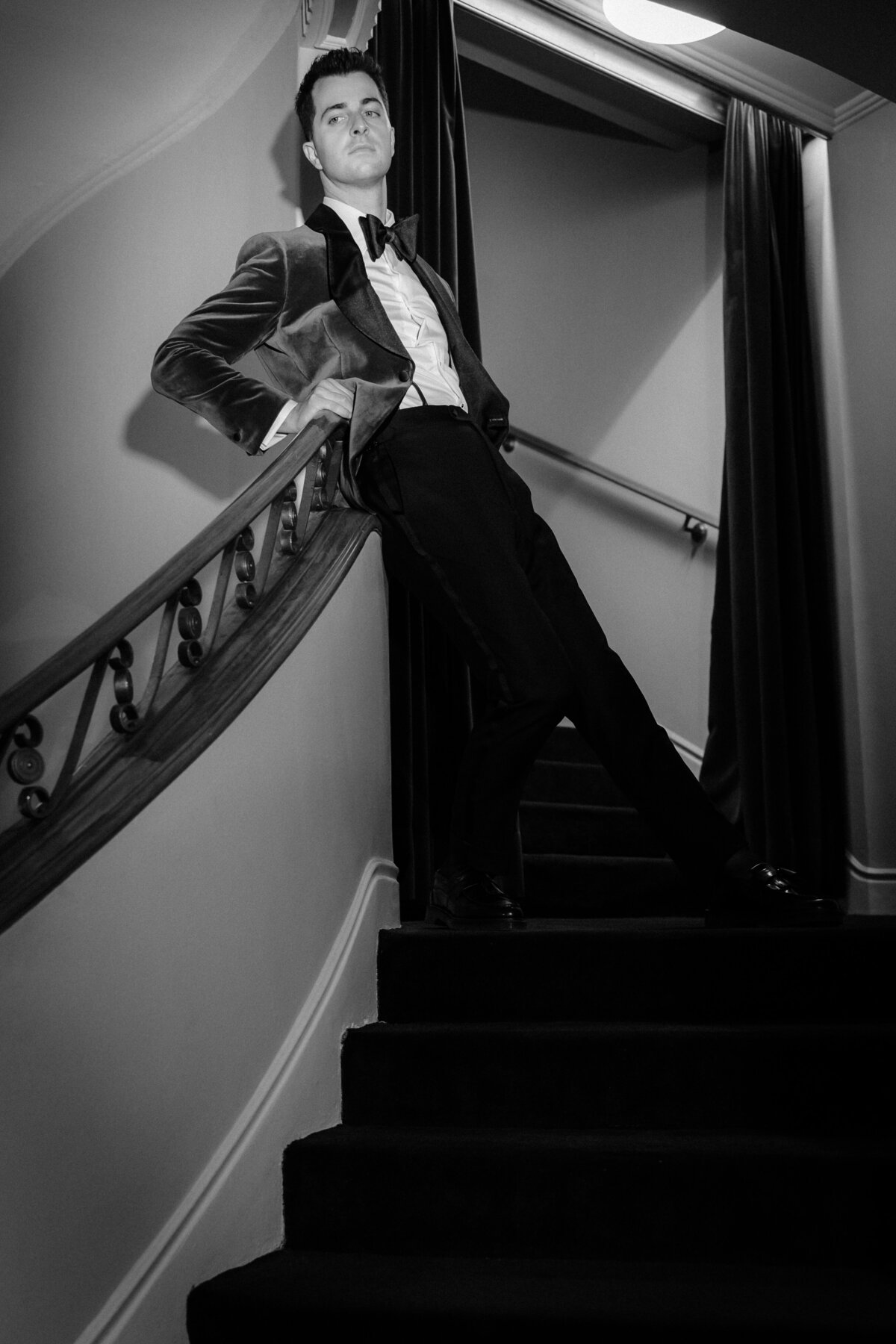 Black and white photo of a groom posing against the hand rail of a staircase.