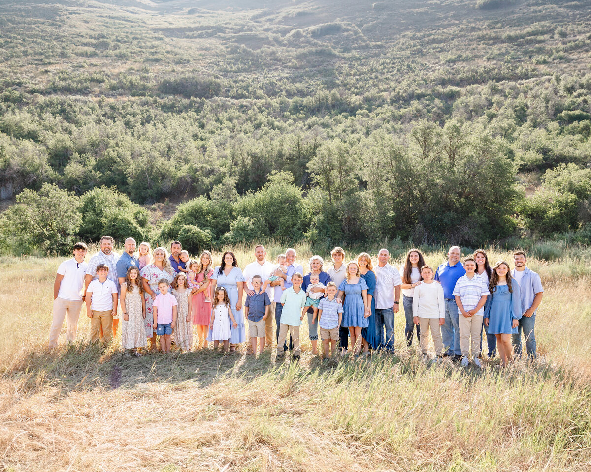 A large family gathers in a picturesque field with a stunning mountain backdrop. Dressed in coordinated pastel and neutral tones, the group celebrates a special moment together, beautifully captured by Cassie Lee Photography.