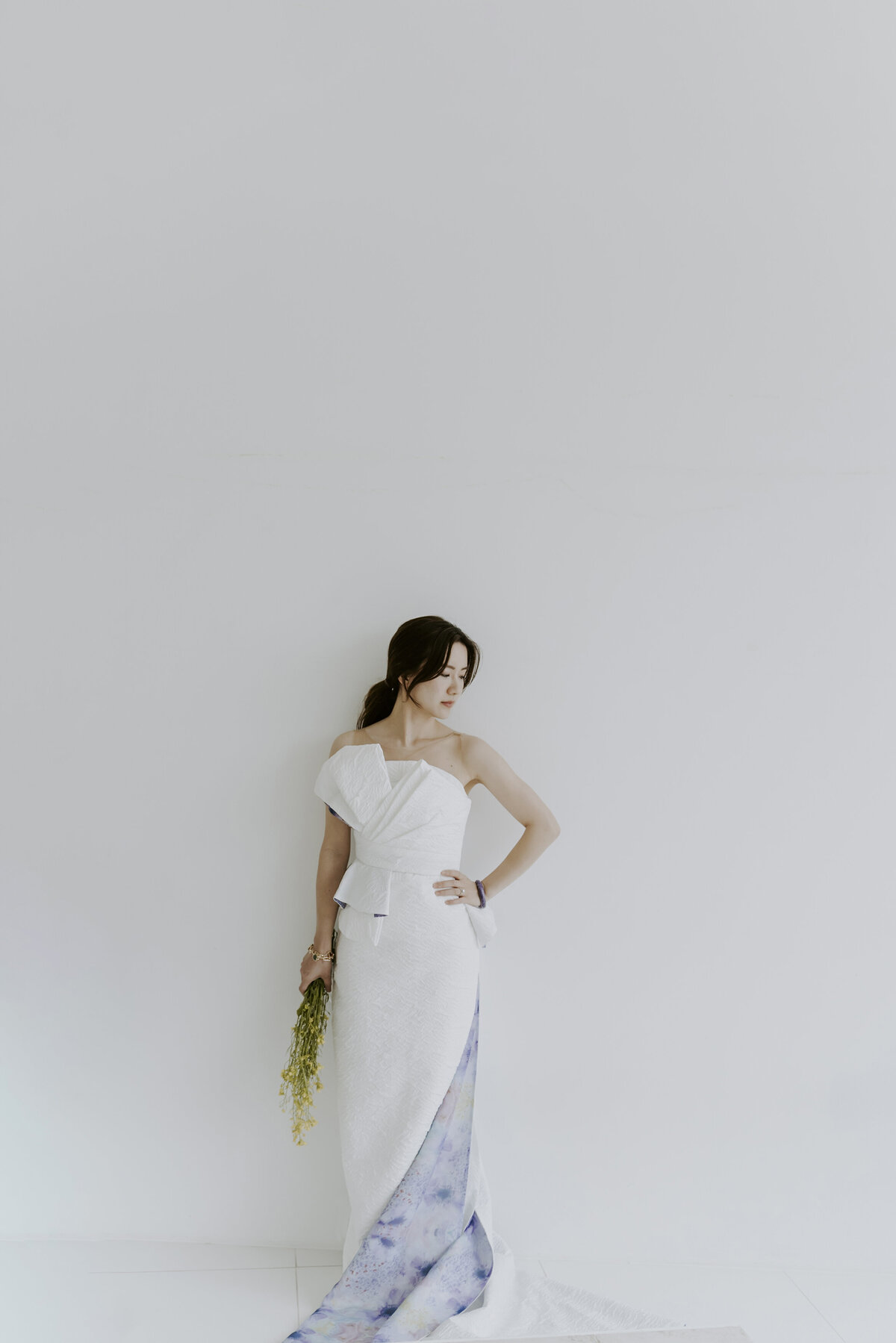 the bride wearing white tube long dress with purple details at the bottom while holding her fresh canola flowers leaning in a white wall