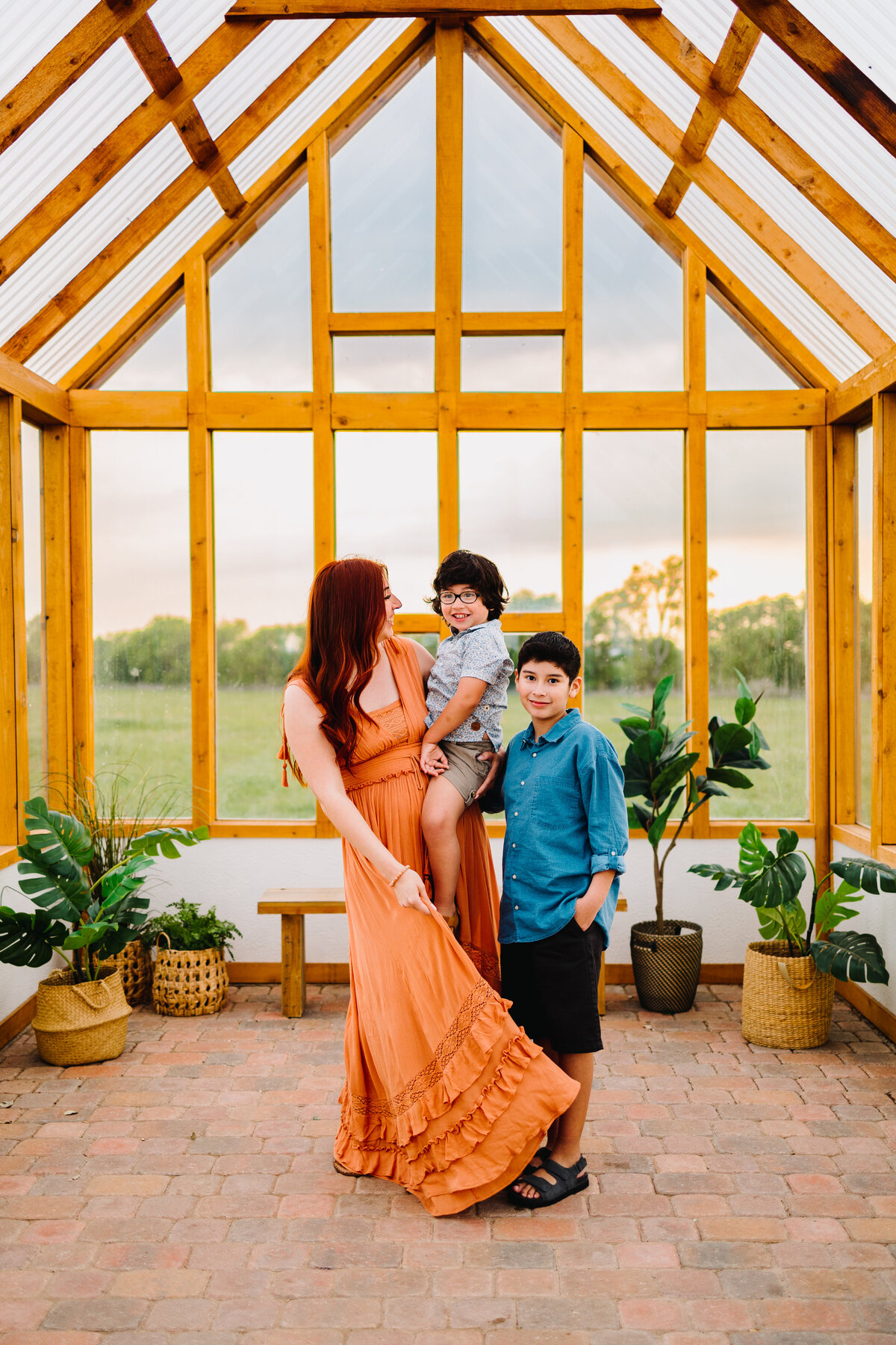 Captured by a professional family photographer in Albuquerque, this photo features a family in a glass cabin surrounded by green plants. The woman in an orange long dress is holding a child wearing glasses and a light blue shirt, while she looks on. The older child, dressed in a dark blue shirt, is smiling at the camera