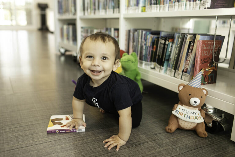 Baby boy in a cute outfit, exploring a stack of books at the library, with a joyful expression during his first birthday photoshoot