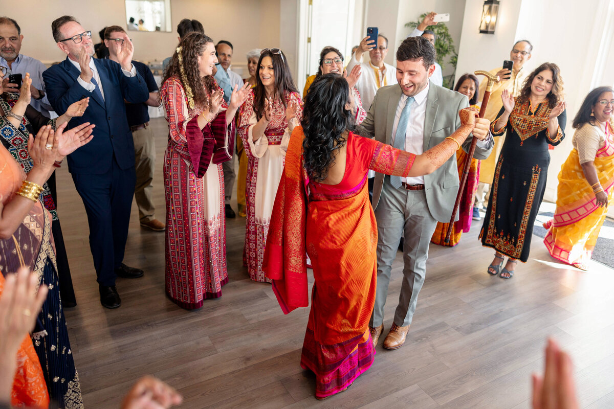 A lively celebration featuring people in colorful traditional attire dancing and clapping. The scene includes men and women, some dressed in suits, surrounded by onlookers capturing the moment on their phones, in a bright and joyful atmosphere.