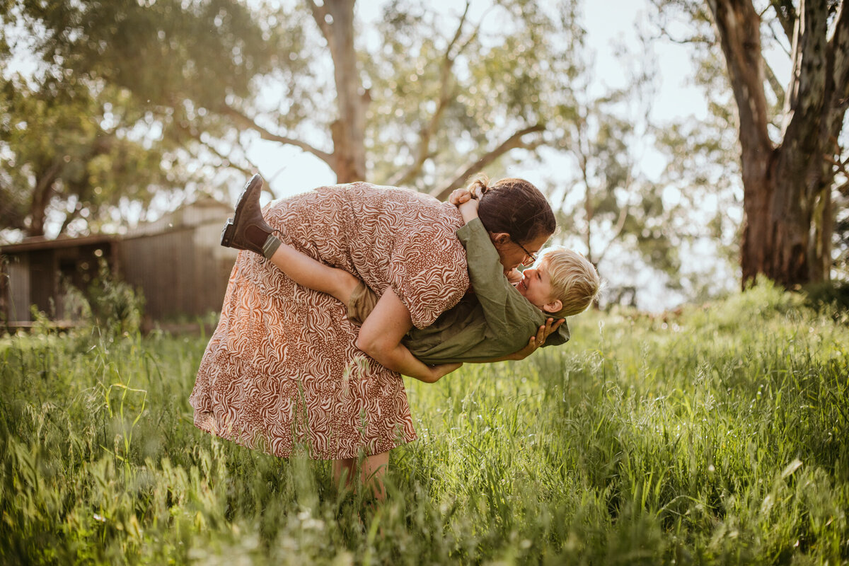 Mom playfully lifts her son in a grassy field, his arms around her neck as he laughs joyfully.