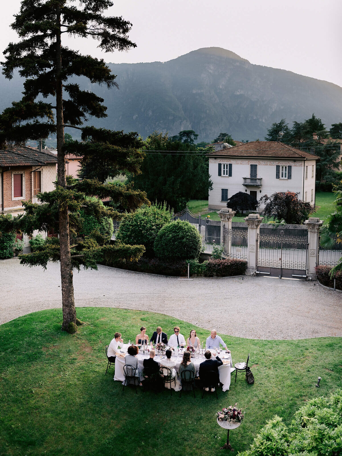 Top view of a group of people on a dining table in a garden, with trees, plants, villa and mountain in the background.