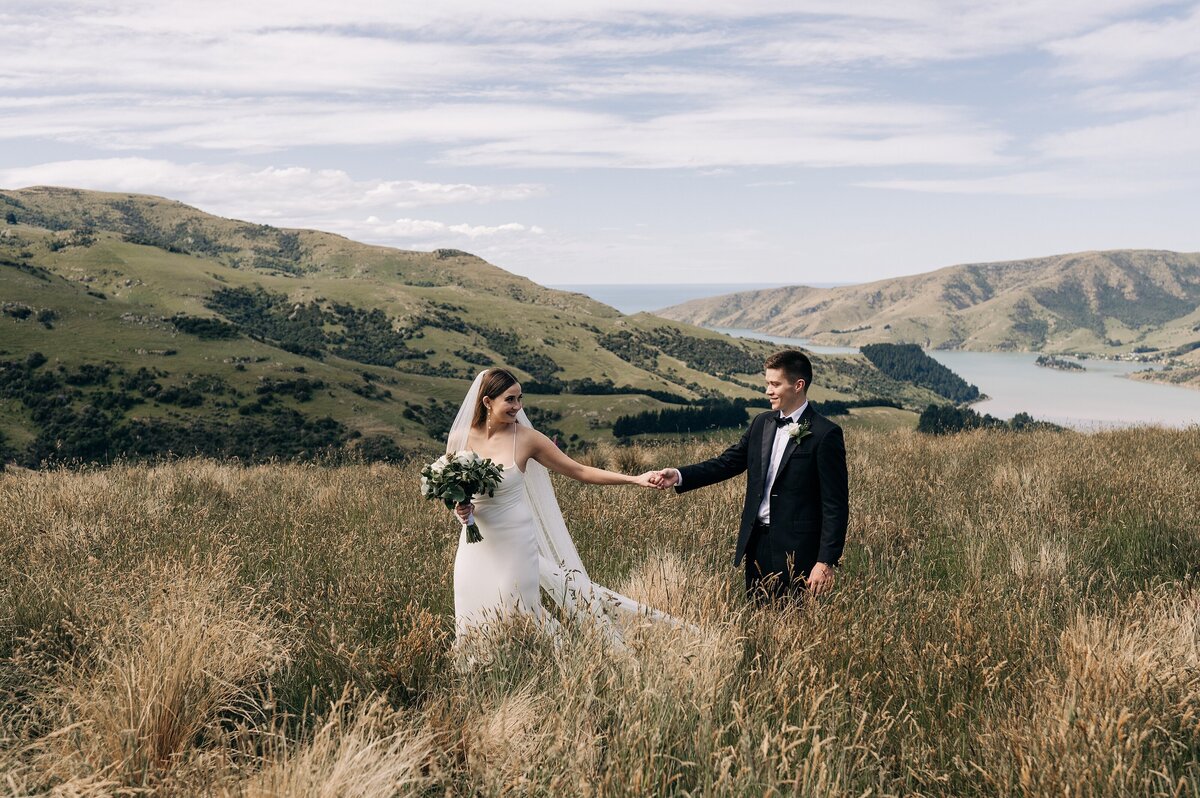 bride in white dress and groom in black suit walking on mountaintop above port levy in new zealand on their wedding day holding hands