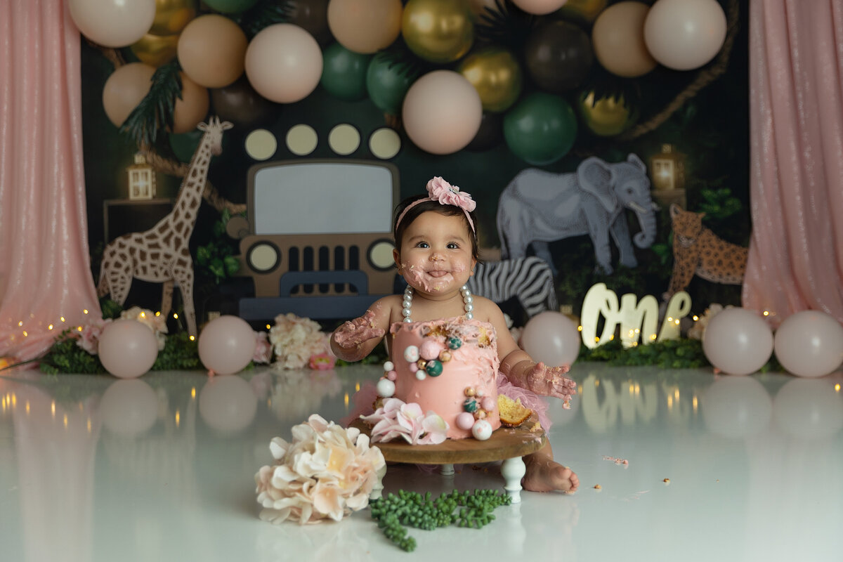 A toddler girl in a pink tutu sits happily covered in cake during her first birthday photo session