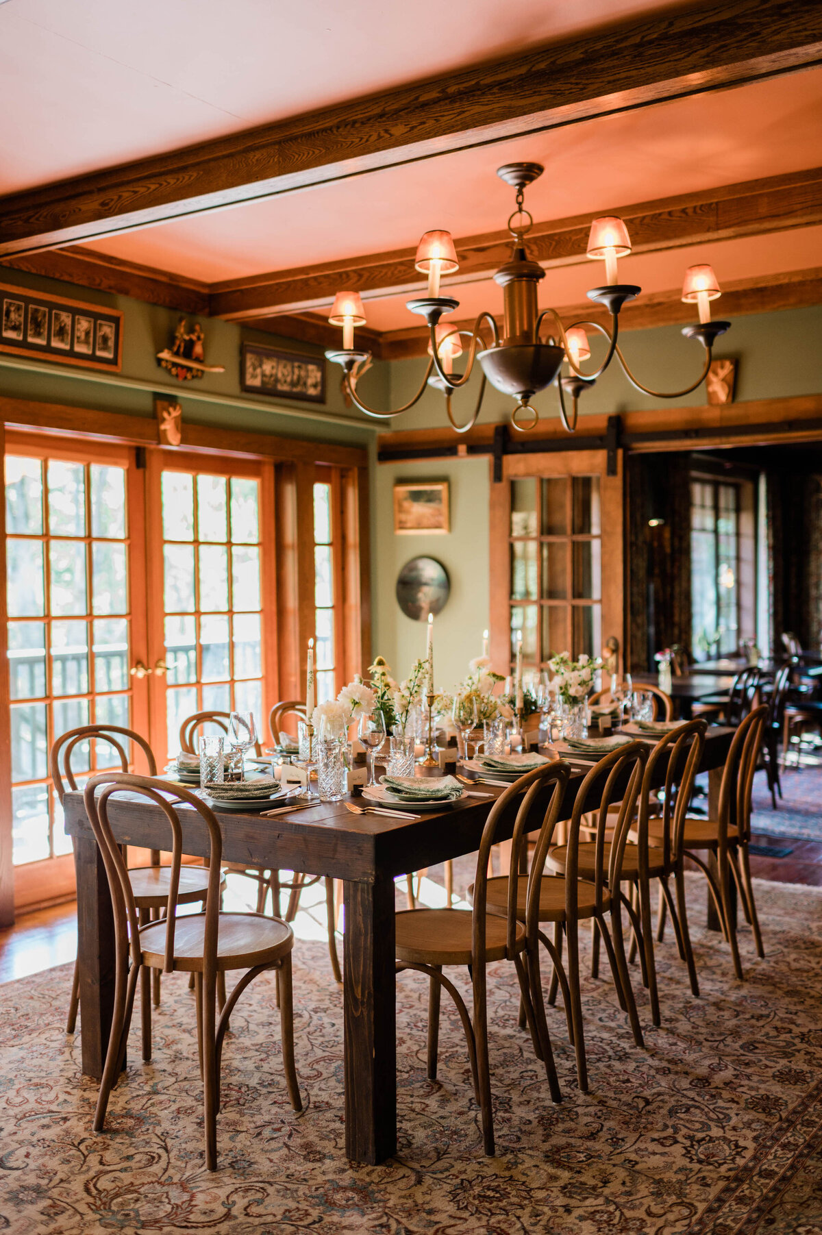 A long wooden dining table inside a warmly lit room is decorated with floral arrangements, candles, and beautifully set place settings with green napkins.