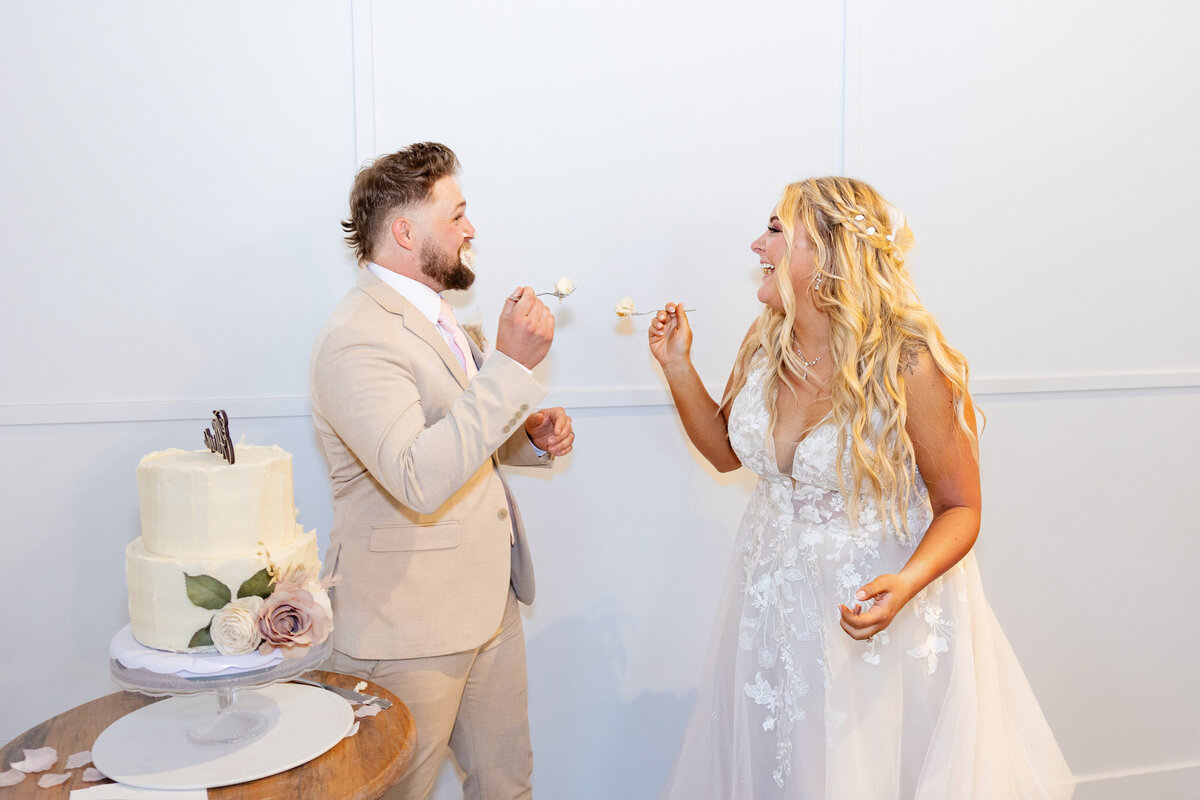 The bride and groom smile and feed each other wedding cake, sharing a playful and sweet moment together.