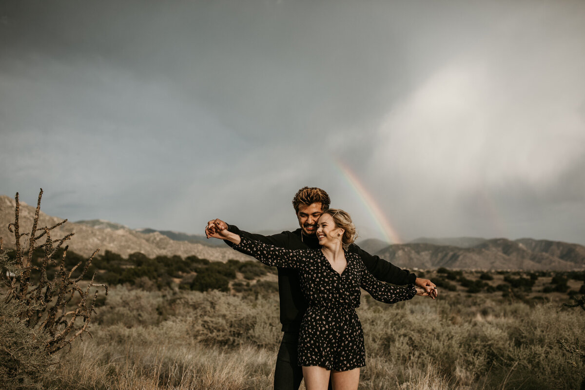 couple holding hands like airplane in front of a rainbow