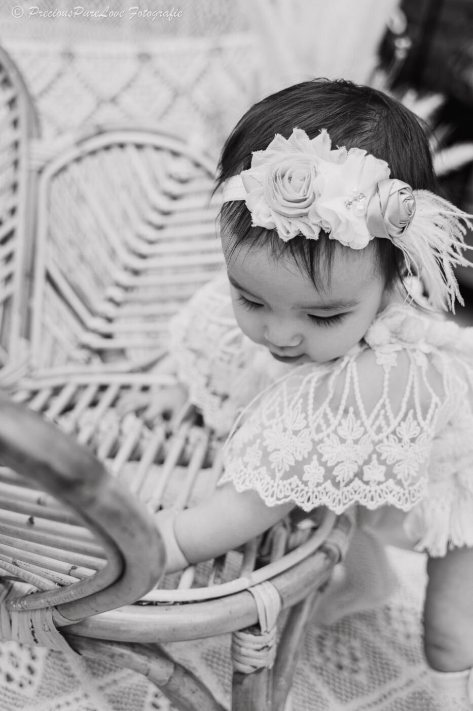 Black-and-white side profile of a baby girl in a lace dress and floral headband, leaning on a wicker chair and looking down.