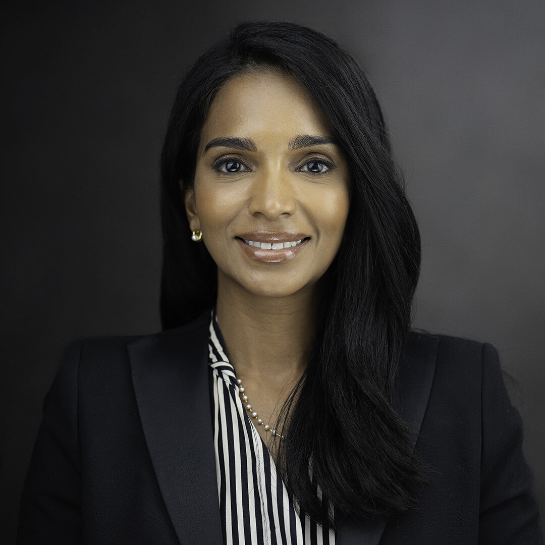 Executive headshot of professional woman in black blazer with striped blouse, confident corporate portrait on dark background by Alex Kaplan Photo.
