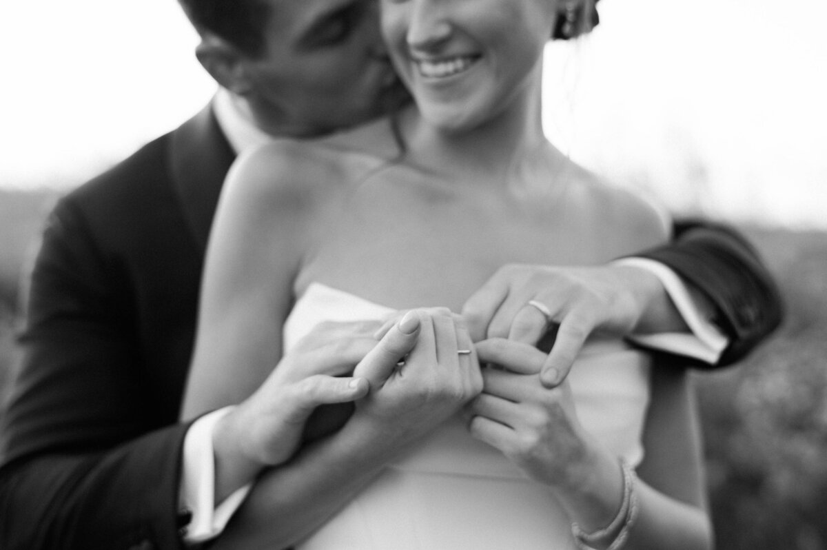 the groom tenderly kissing the bride’s shoulder while holding her hands