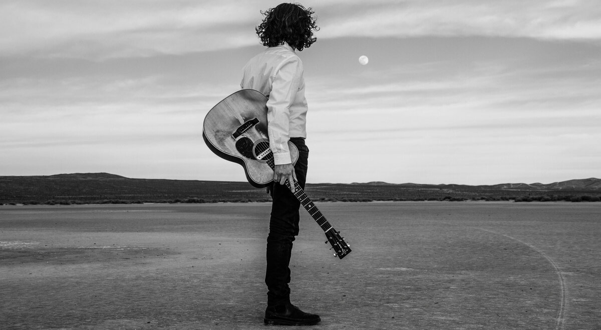 Musician portrait black and white Noah Zacharin standing in desert back to camera looking sunset while holding acoustic guitar El Mirgae