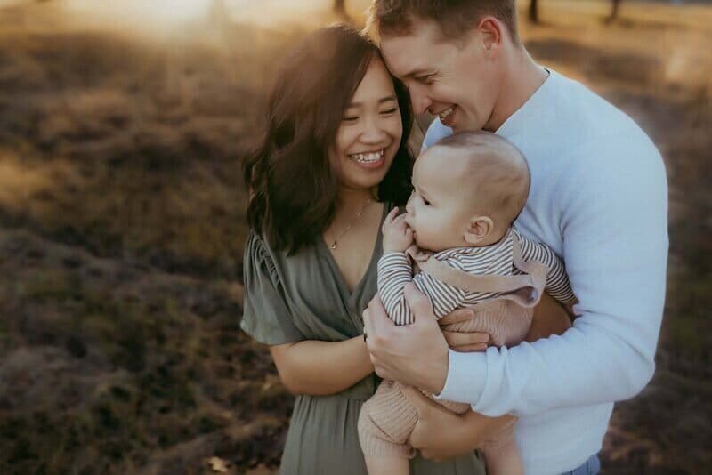 A couple stands closely together in a field, smiling and holding a baby. The sunlight creates a warm and soft atmosphere, perfectly captured by a family photographer in Warner Robin GA. 