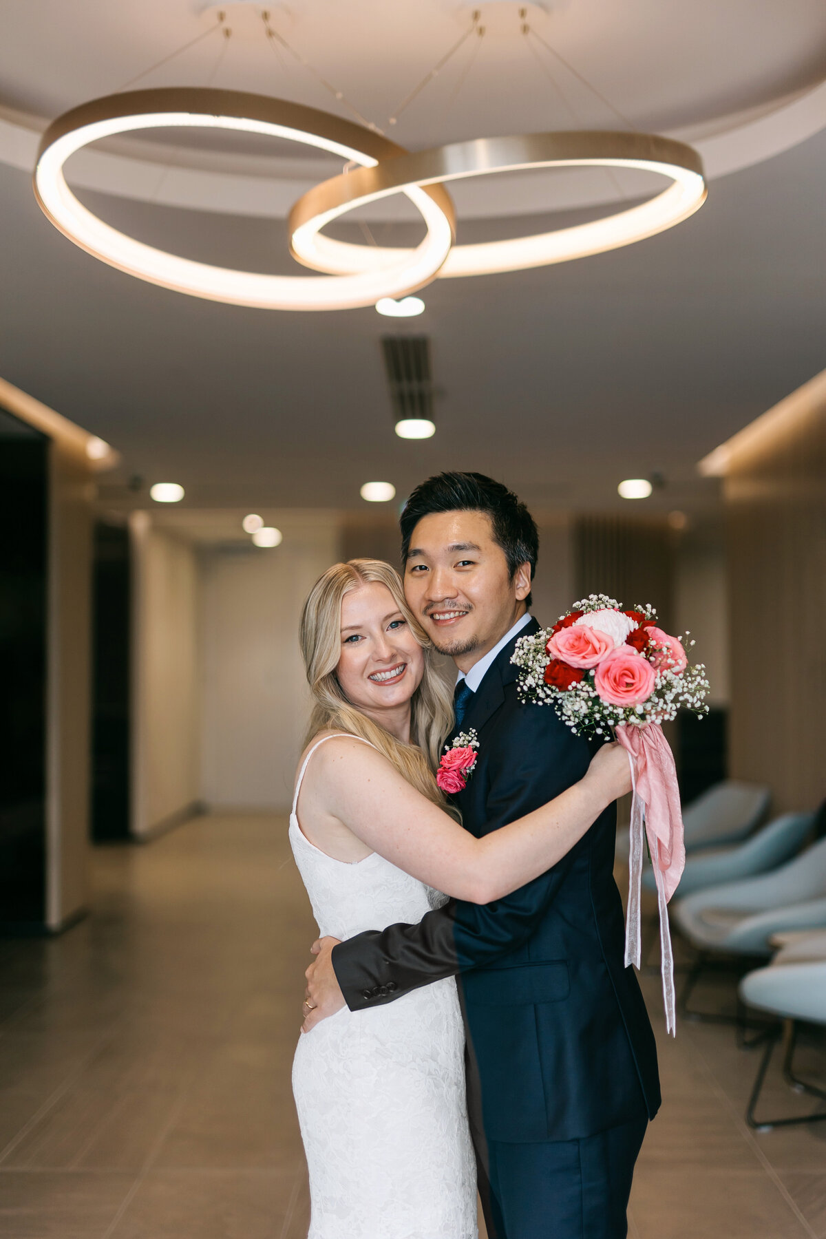 Registry Office bride and groom posing indoors beneath a modern circular chandelier, smiling after their ceremony.