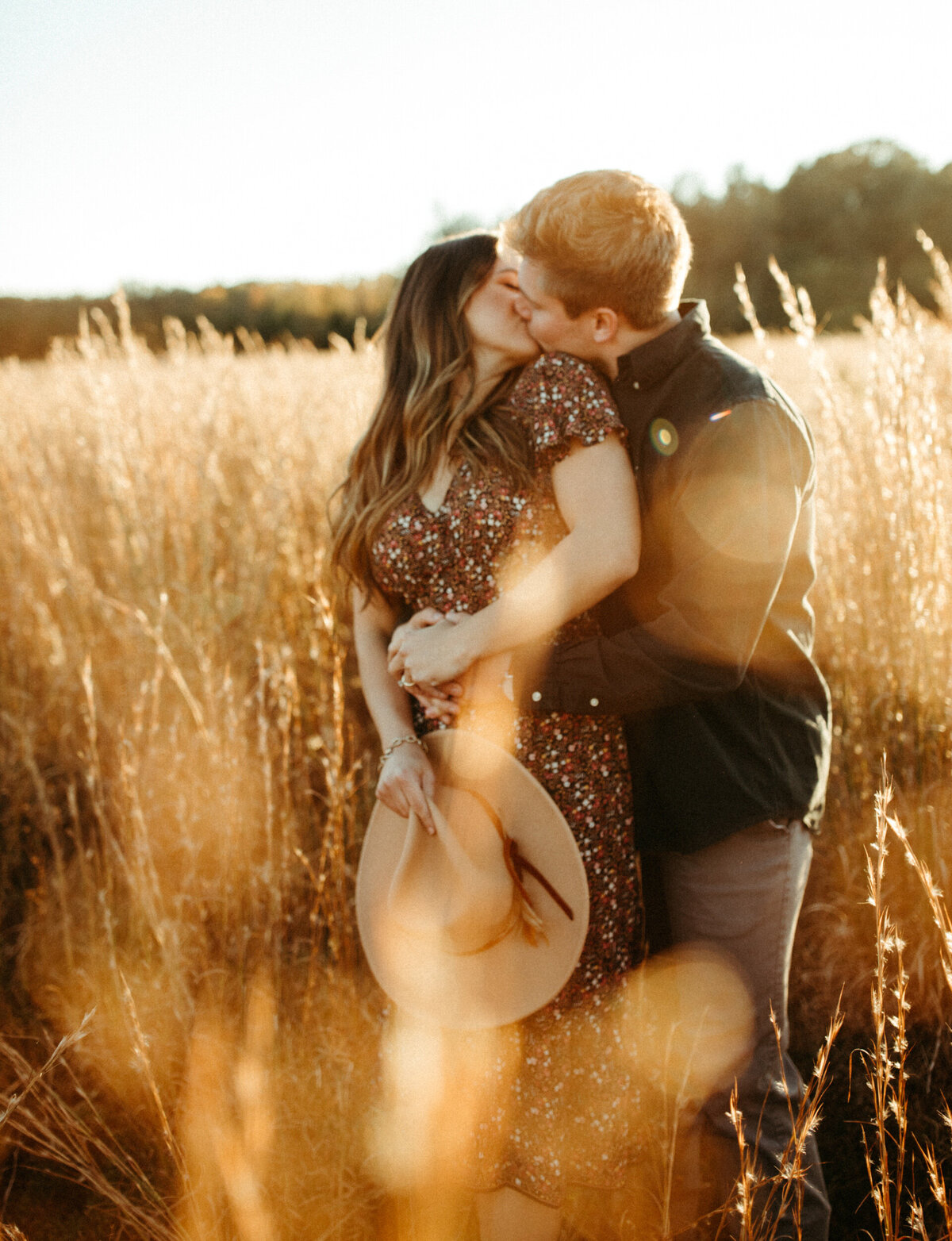 Girl wearing floral dress and holding boho hat is kissing her fiancé while he stands behind her and wraps his arms around her in a field with tall golden grass