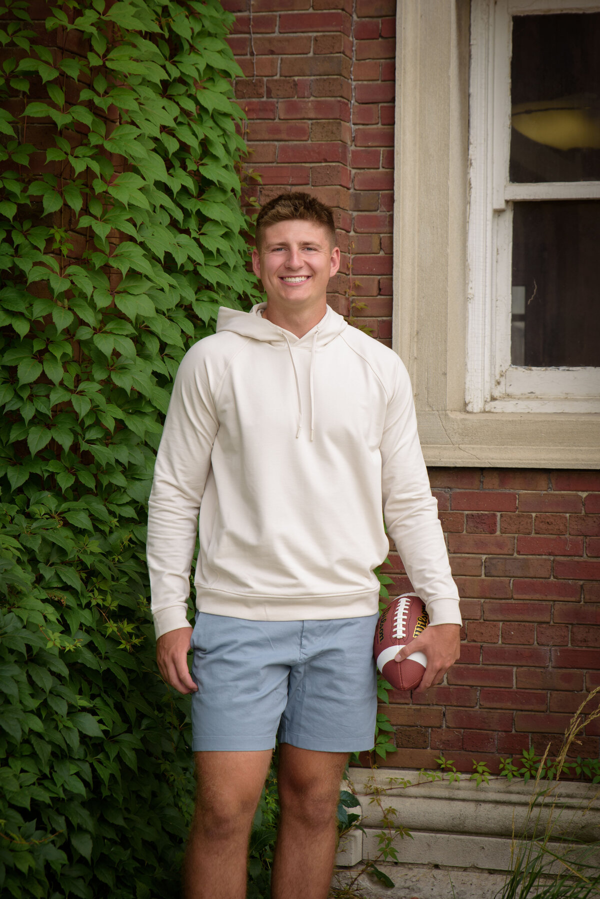 De Pere High School senior boy wearing blue shorts and a cream hooded sweatshirt standing holding football in an urban setting at The Depot in Downtown Green Bay, Wisconsin