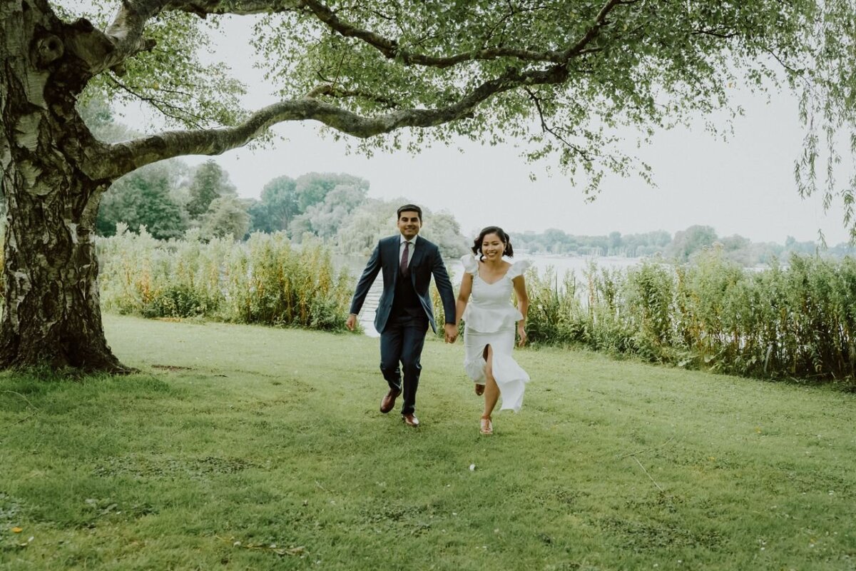  A cheerful candid photo of a couple running towards the camera on a grassy lawn. The man, in a dark blue suit, and the woman, in a white dress with a slit, are holding hands and smiling as they run. A large, twisting tree is on the left, and tall grass and a body of water are in the background under a soft, cloudy sky.