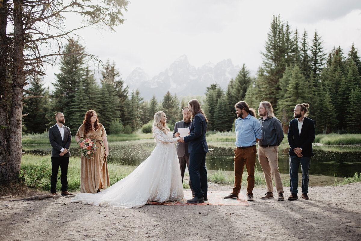 schwabacher landing wedding ceremony with bride and groom holding hands while standing next to a river with their friends and family for their outdoor ceremony with a view of the Tetons